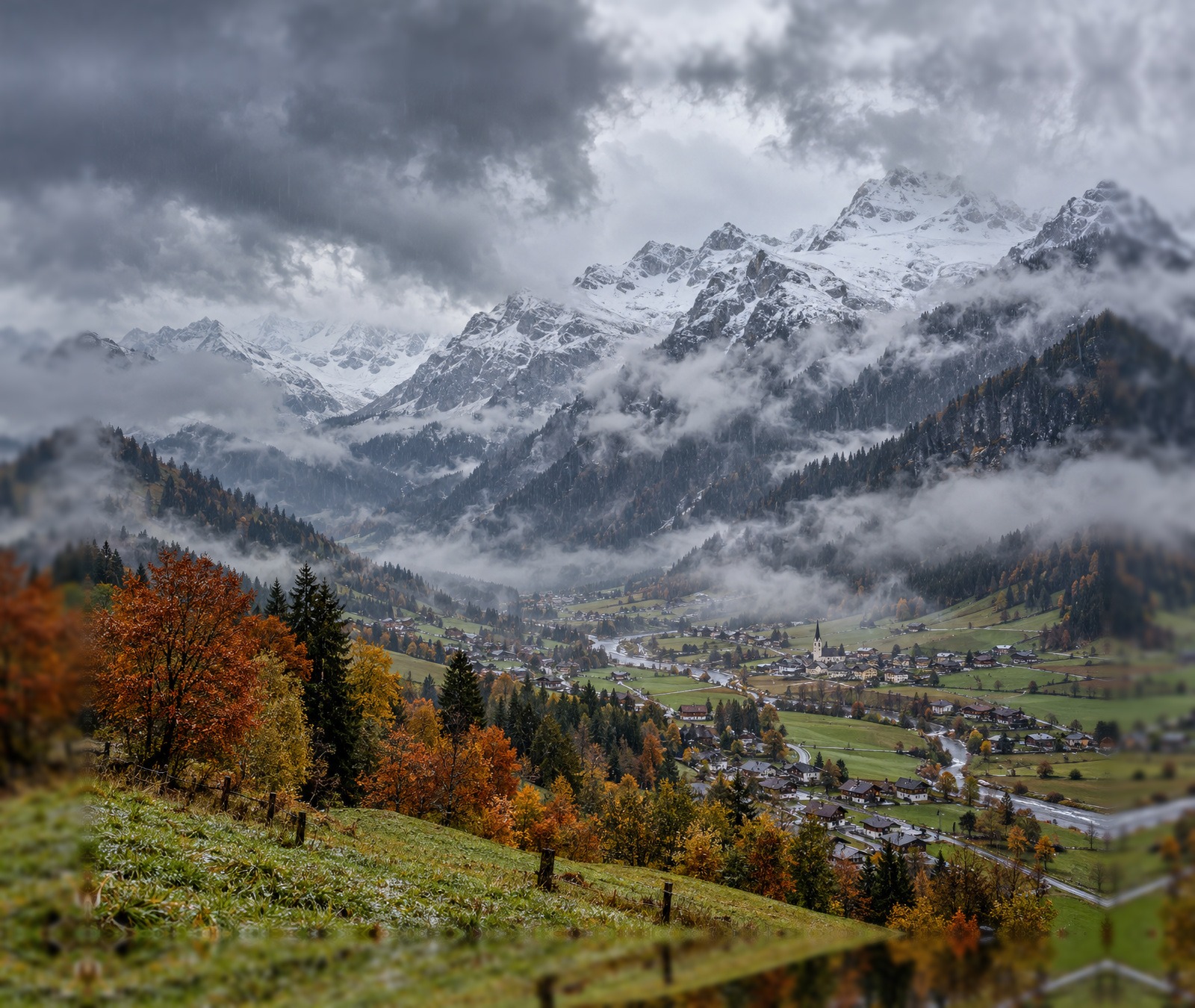 Herbstliches österreichisches Alpental unter dunklen Kaltfrontwolken mit frischem Schnee auf den Gipfeln.